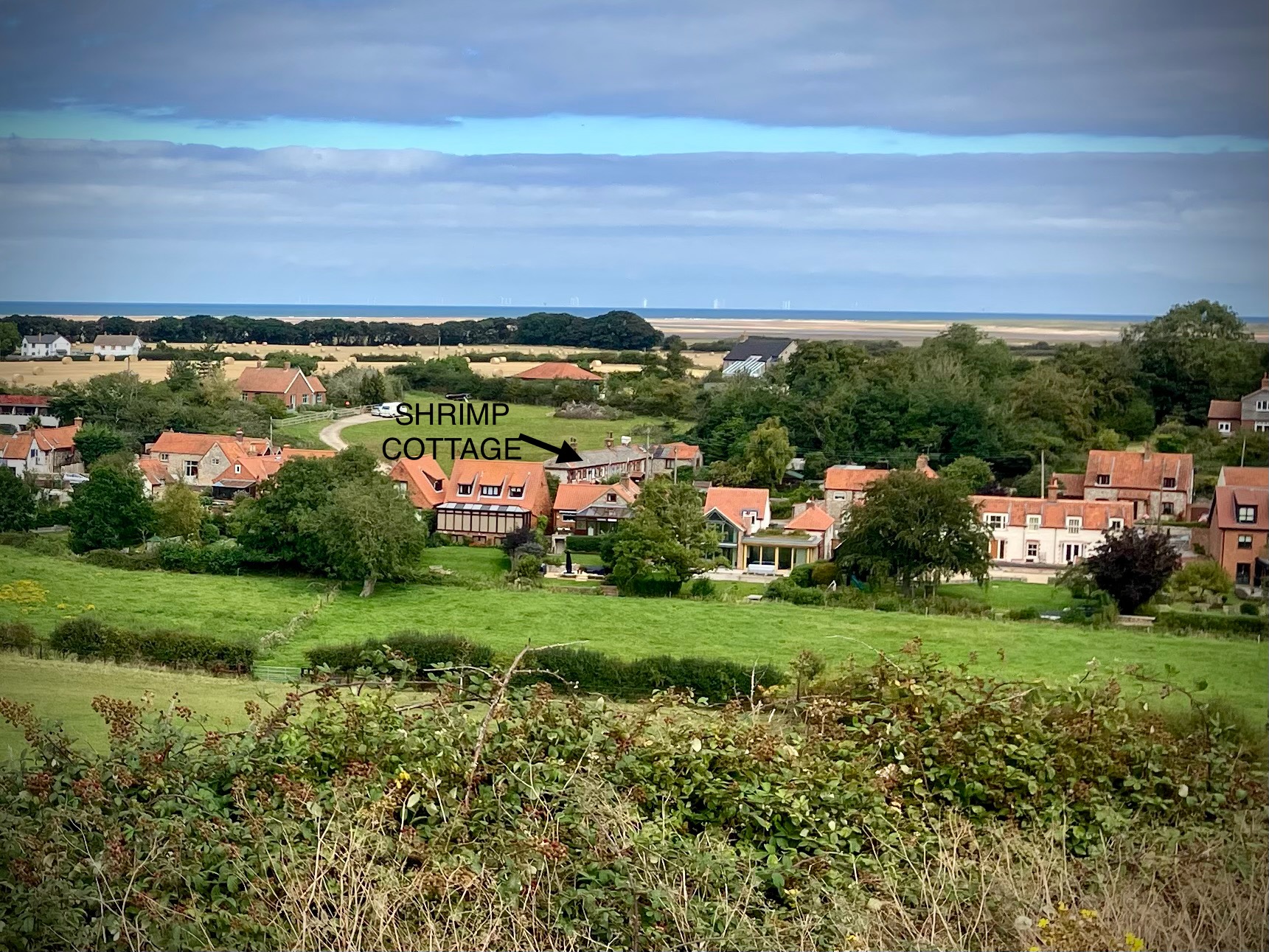 Countryside village view with green fields, red-roof cottages, distant sea, and arrow pointing to Shrimp Cottage in Stiffkey Norfolk.